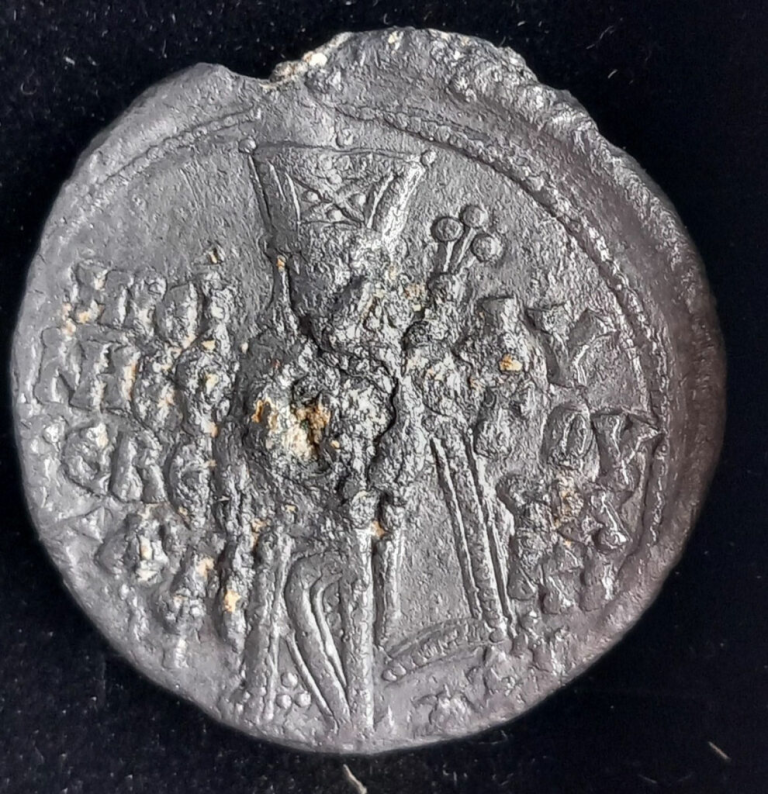 Ancient circular coin with a standing figure in the center, holding a tall staff or scepter, surrounded by worn inscriptions and decorative marks on a rough, pitted surface.