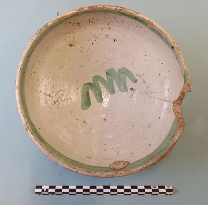 Round ceramic bowl with a pale interior, green rim, and a green spiral mark inside; chipped edge and a measurement scale beneath on a teal surface.
