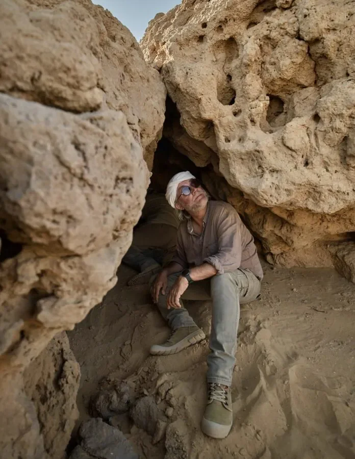 Cerveteri-Egitto Man with a white headscarf and sunglasses sits in a sandy rock alcove, leaning against a rough cave wall.