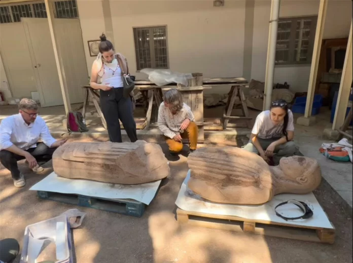 Colosso di Sardi Four artists in a courtyard studio examine two large stone busts resting on pallets outside a workshop.