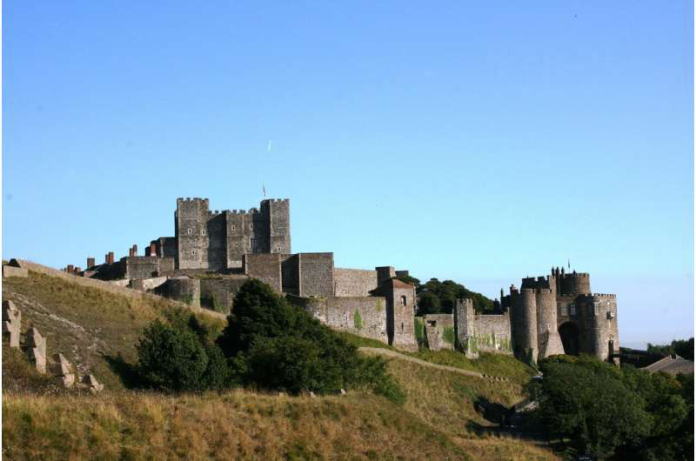 Medieval stone castle atop a grassy hill with towers and surrounding walls under a clear blue sky.