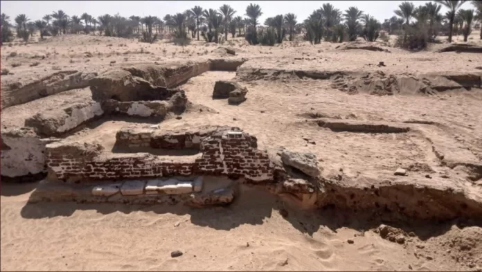 Desert archaeological site with partially preserved brick walls and sun-baked soil, palm trees in the distance.