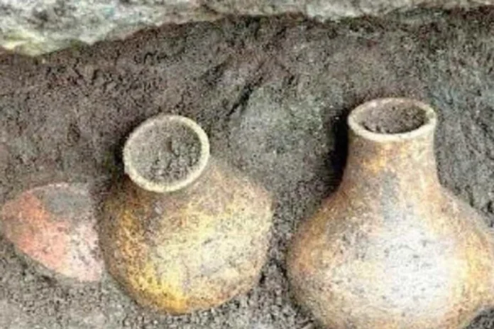 Two weathered clay pots partially buried in soil, one with a short neck and wide opening and the other with a taller narrow neck, both dusty and worn.