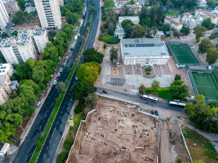 Aerial view of a city block: tree-lined road, a large building with parking, adjacent sports fields, and an excavation site in the foreground.