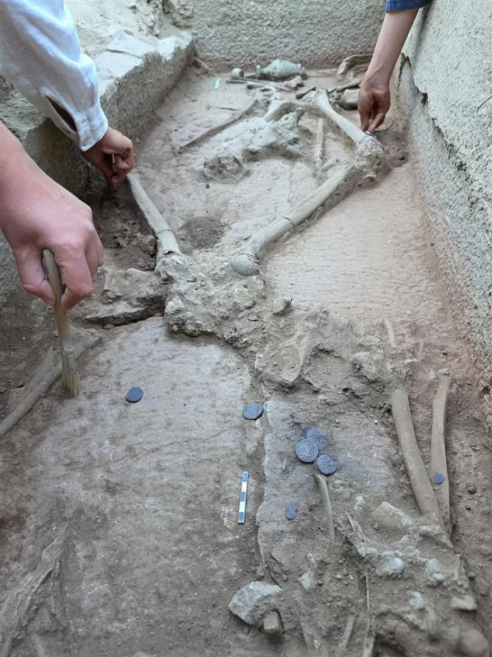 Lottatore Archaeologists brushing dirt in a trench, uncovering a partial skeleton with bones, coins, and a measuring scale nearby.