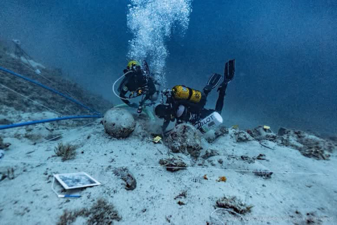 Two divers in scuba gear operate an underwater remotely operated vehicle on a sandy seabed, with tether lines and rising bubbles.