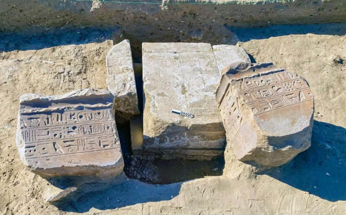 Archaeological excavation with three large stone blocks carved with hieroglyphs partially buried in sandy ground.