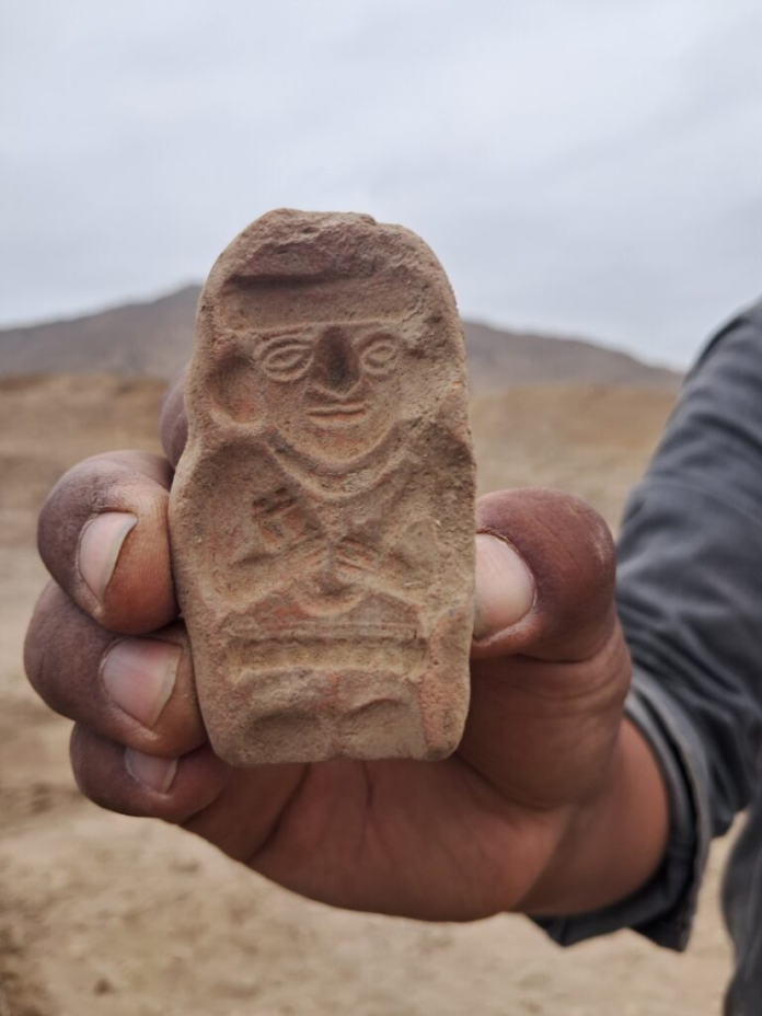 Hand holding a small carved clay figure with a smiling face in a desert landscape.