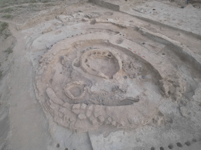Archaeological site with concentric circular trenches and a central pit, surrounded by scattered stones and soil.