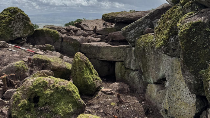 Rugged, mossy rock terrain with stacked boulders and a dark crevice at the center under a cloudy sky.