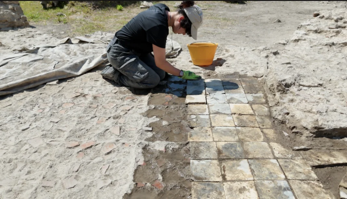 Person kneeling in a dusty outdoor area, cleaning square tiles to form a paved path with a yellow bucket nearby.