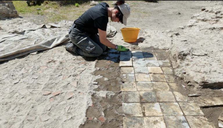 Person kneeling in a dusty outdoor area, cleaning square tiles to form a paved path with a yellow bucket nearby.