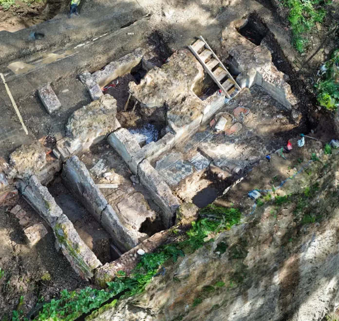 Aerial view of an archaeological excavation with exposed stone walls, trenches, and a wooden ladder. It shows researchers at a historic ruin site on a hillside.
