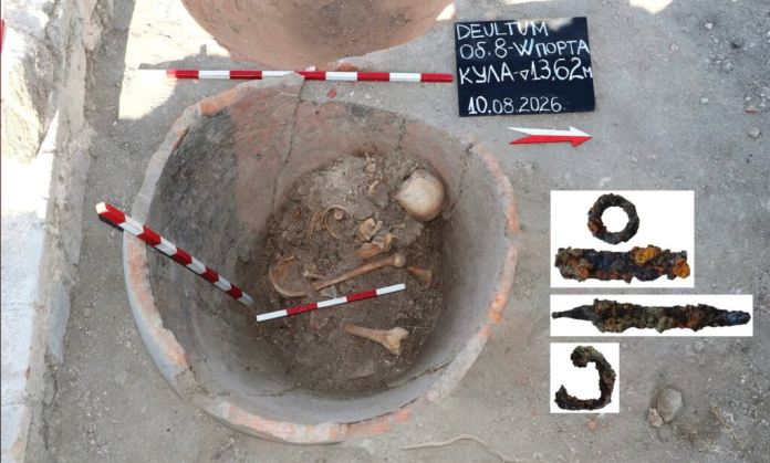 Bulgaria_Dolium_Burial_Knives Open excavation pit with bones in soil, red-and-white measuring rods, and a chalkboard with site details; inset shows three corroded metal artifacts (ring and rods).