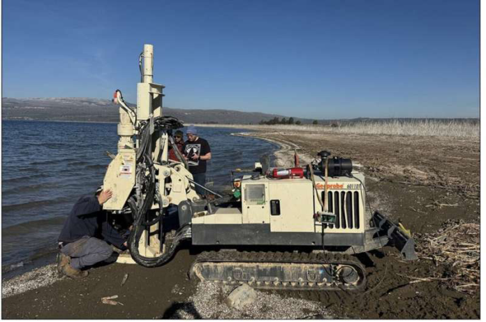 A compact tracked drilling rig on a sandy beach beside calm water, with two workers inspecting the controls.