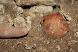Ceramica Close-up of a finger beside a small red clay ball partially buried in dirt.