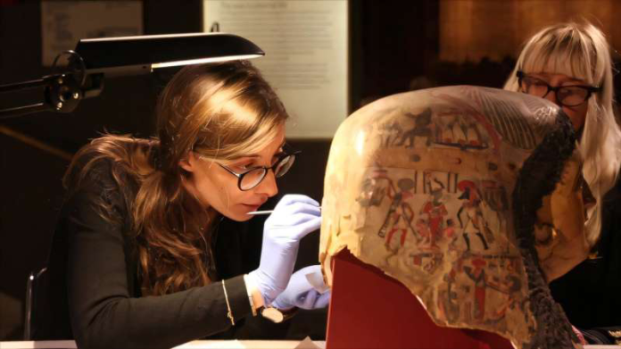 Conservator in gloves examines an ancient decorated mummy head with precision tools in a lab setting.