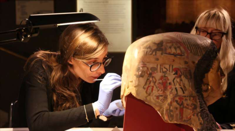 Conservator in gloves examines an ancient decorated mummy head with precision tools in a lab setting.