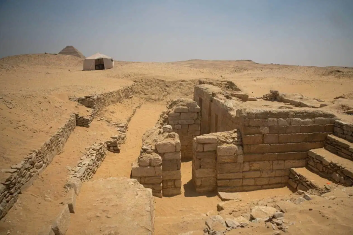 Sunlit desert ruins with stone walls and stair steps in the sand, a white shelter in the distance, and a pyramid on the horizon.