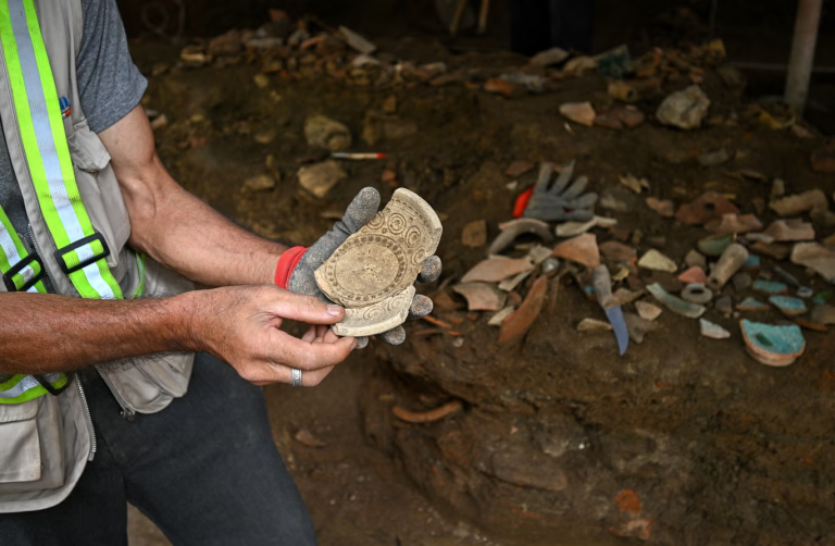 Worker in a reflective vest holds two decorated ancient ceramic shard fragments at a dirt excavation site