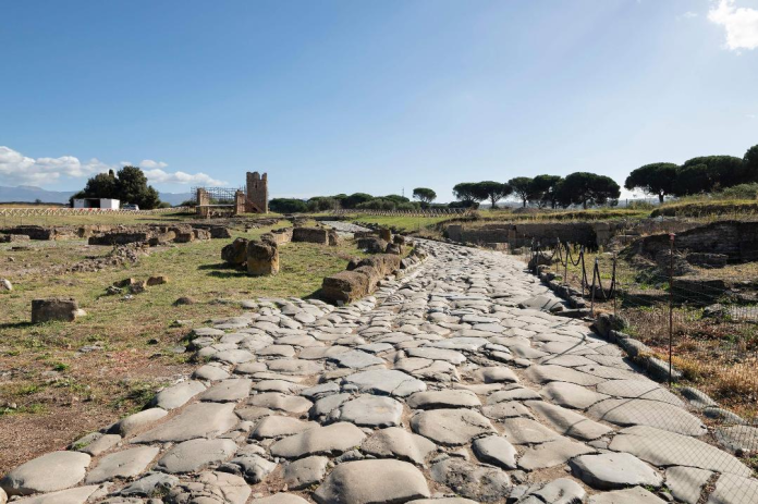 Stone cobblestone path winds through ancient ruins toward a distant tower under a clear blue sky.