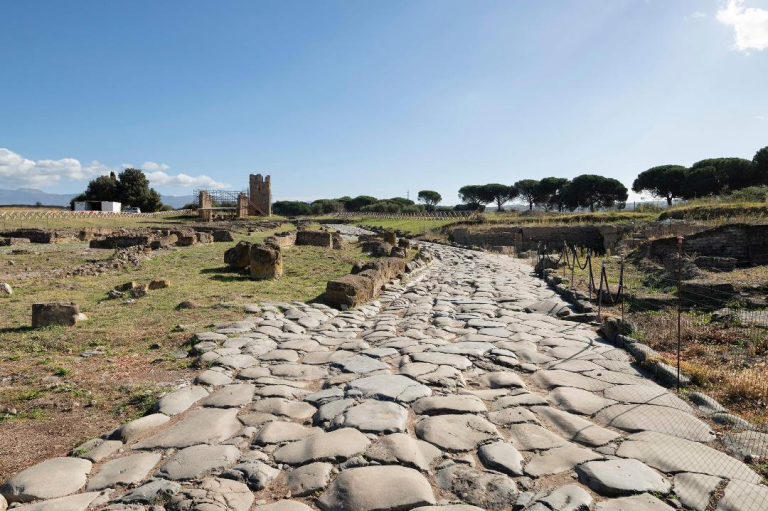 Stone cobblestone path winds through ancient ruins toward a distant tower under a clear blue sky.