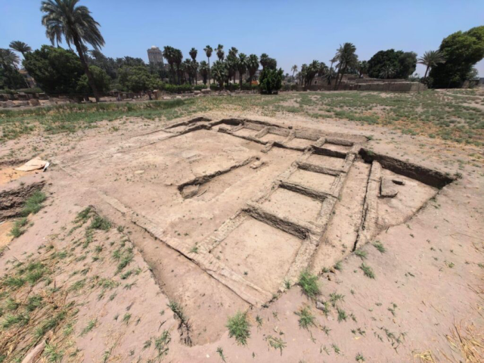 Menfi_Mit_Rahina Archaeological trench with rectangular stone foundations exposed on a dusty site, palm trees in the background.