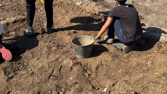 Navalia People working at a dirt construction site; one person kneels beside a metal bucket in a small excavation area.