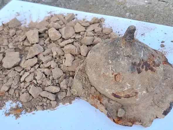 Scudo Vandalo Old weathered clay pot partially buried in dirt with a pile of small rocks nearby on a white surface.