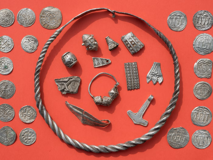 Tesori Vichinghi Assorted ancient metal jewelry and coins arranged on a red background, inside a twisted rope circle.