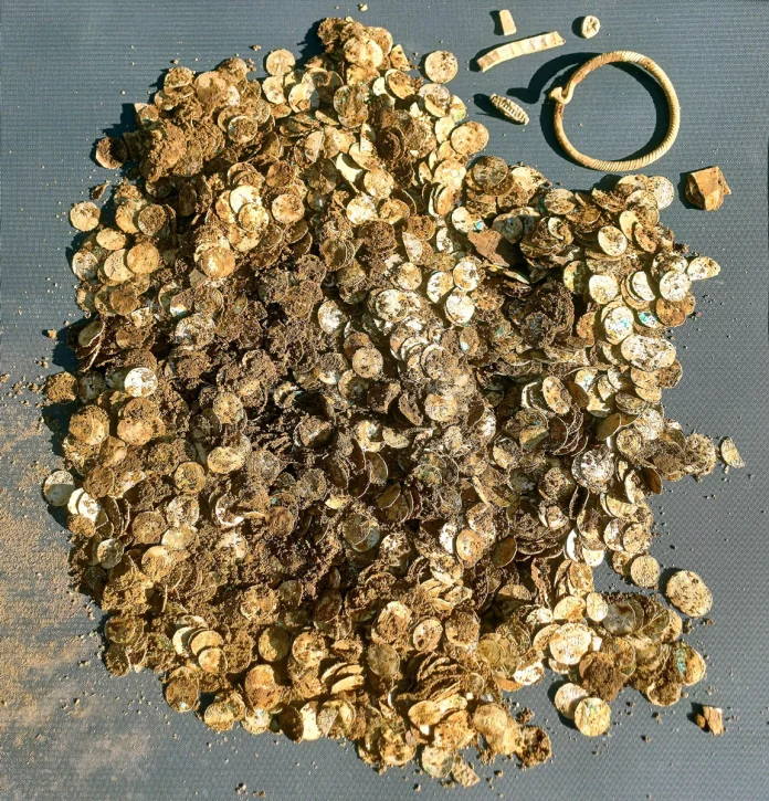 Tesoro_medievale Heap of dusty gold coins covered in dirt on a gray surface, with a circular bracelet to the right.