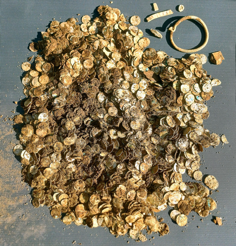 Heap of dusty gold coins covered in dirt on a gray surface, with a circular bracelet to the right.
