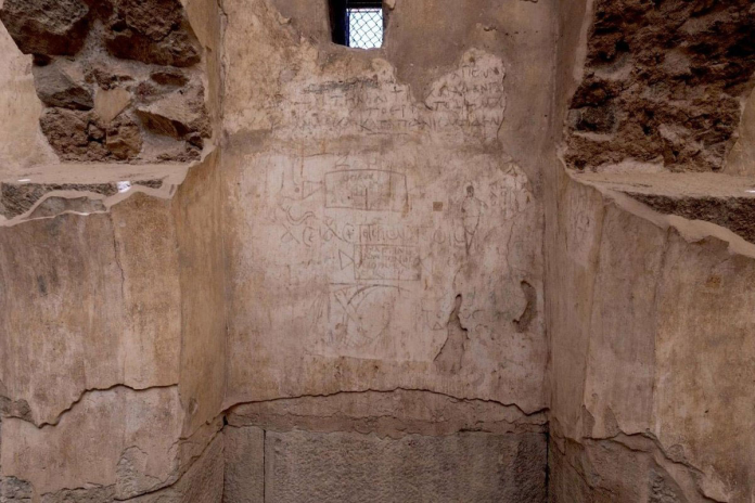 Interior view of an old stone-walled room with plaster, showing graffiti scribbles on the wall and a small barred window near the ceiling.