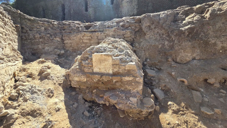 Ruins with rough stone walls and a central mound containing a rectangular stone block, dirt and rubble around the site