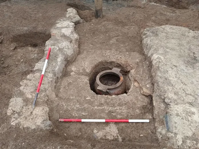 pontecagnano-tomba Circular rusted pipe flange recessed in a trench, with exposed earth and stone walls, and red-and-white measurement rods for scale nearby.