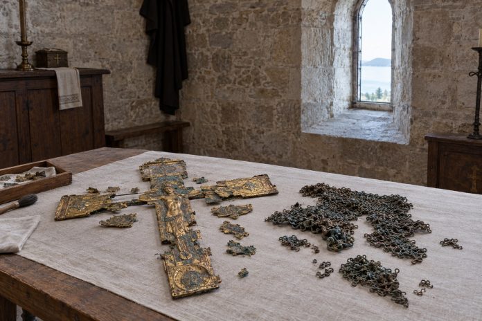 Stone chapel interior with a large ornate crucifix and broken panels laid on a cloth-covered wooden table; piles of metal chains nearby.